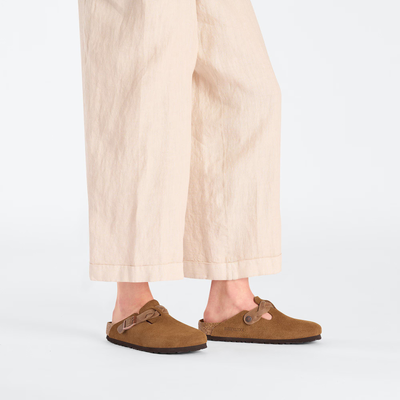 Person wearing Birkenstock Boston clogs in mink suede with a braided strap, paired with cream wide-leg pants.