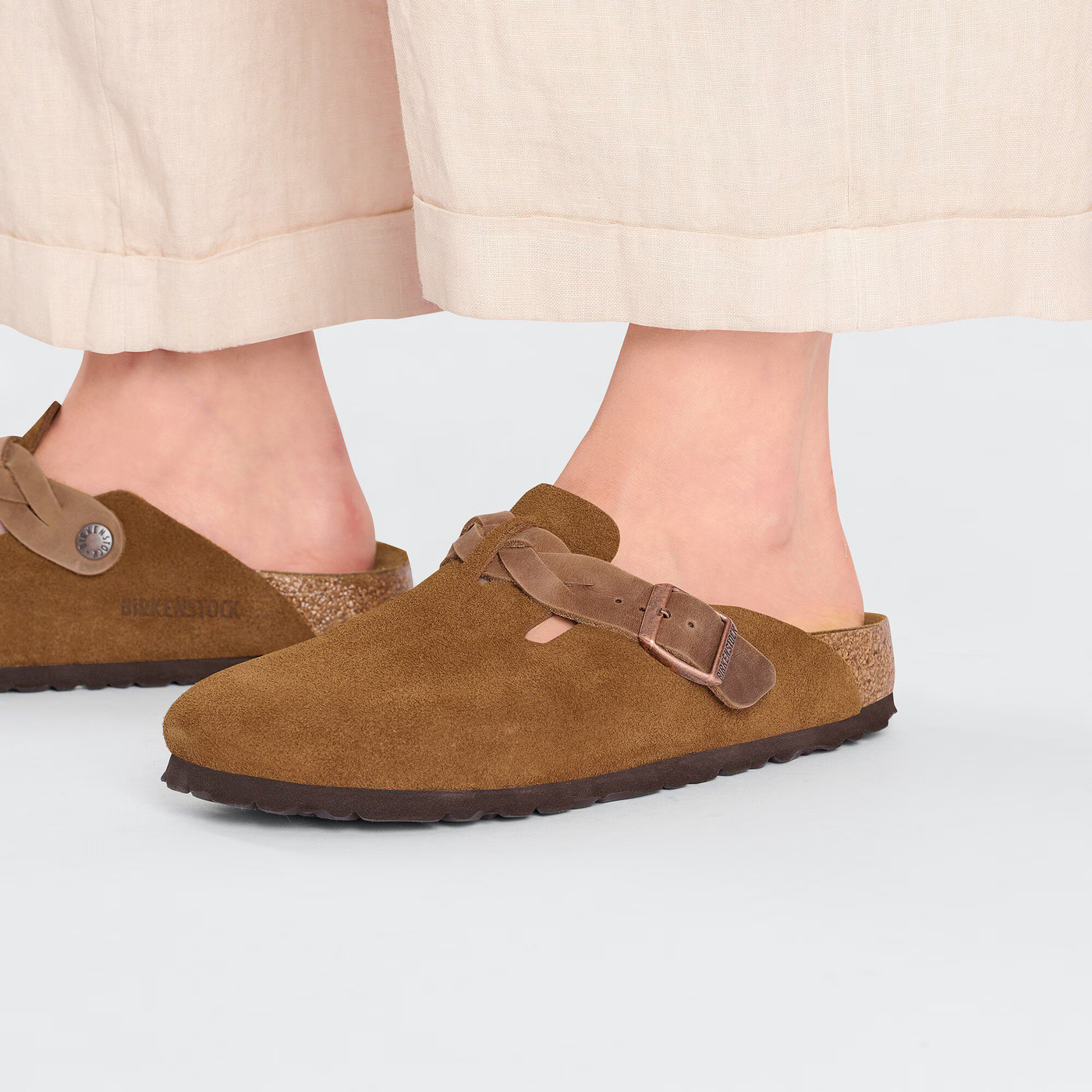 Woman wearing Birkenstock Boston clogs in mink suede with a braided strap, paired with cream wide-leg pants.