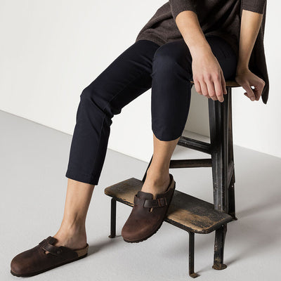 A person seated on a rustic stool wearing dark navy pants and a brown top, styled with Boston Habana clogs in oiled leather, showcasing a casual yet sophisticated look.