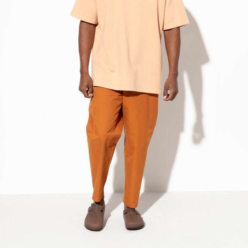 A person standing against a white background wearing a beige oversized T-shirt, rust-colored pants, and Boston Habana clogs, styled for a minimalist and relaxed look.