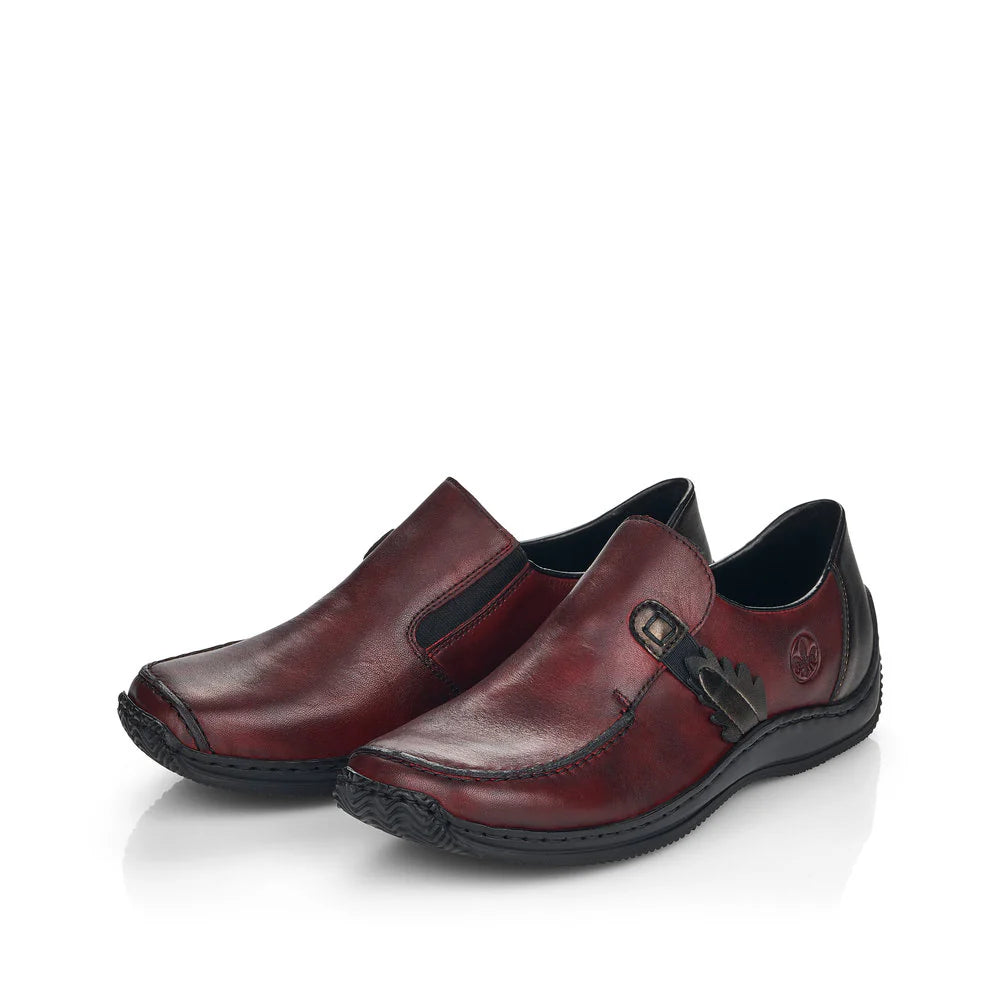Rieker Women’s Burgundy leather slip-on shoes with side elastic panels, decorative strap detail, and flexible black outsole for everyday comfort.
