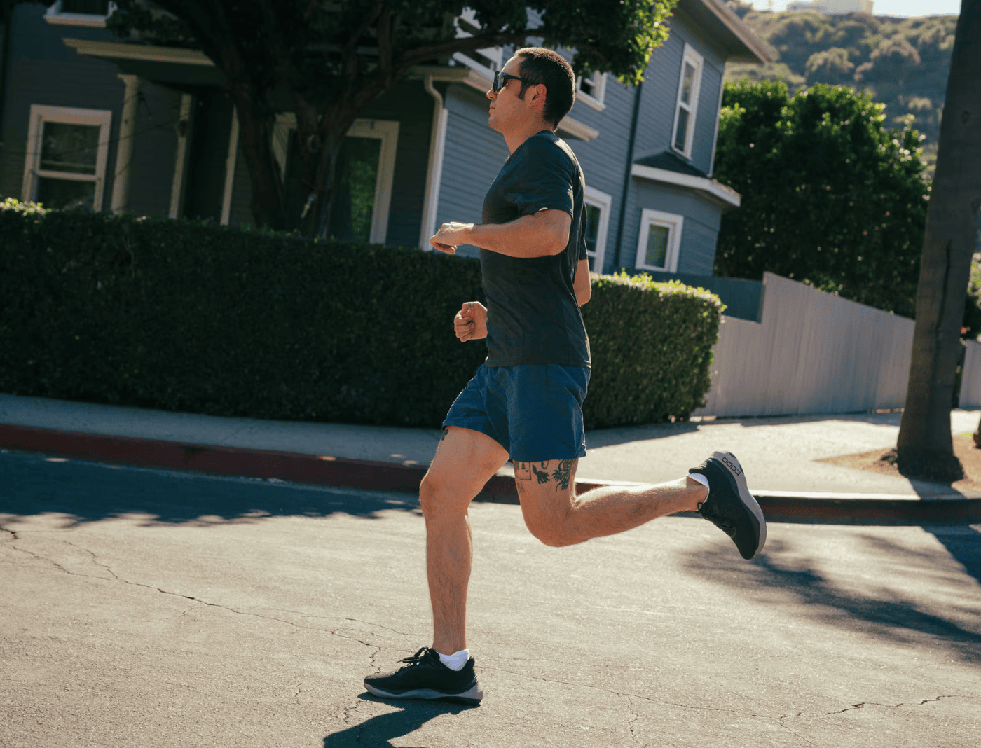 A man running on a sunny residential street wearing all-black Topo running shoes with a gray cushioned sole, blue shorts, and a dark t-shirt, captured mid-stride against a backdrop of houses and greenery