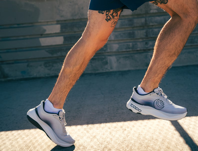 A runner wearing light gray Topo running shoes with a gradient design fading into charcoal, a black swirl logo on the side, and 'topo' branding on the sole, captured mid-stride on a sunlit urban pavement.